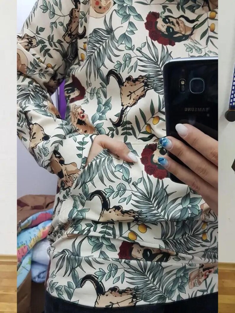 May include: A long-sleeve hoodie in an off-white color featuring a repeating pattern of green tropical leaves, yellow citrus fruits, and stylized skulls adorned with red floral crowns. The garment includes a front kangaroo pocket and ribbed cuffs.