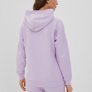 May include: A light purple hooded sweatshirt featuring long sleeves with ribbed cuffs, a matching ribbed hem, and a hood. The garment is constructed from a solid-colored fabric and is shown from the back, displaying the hood and sleeve seams.