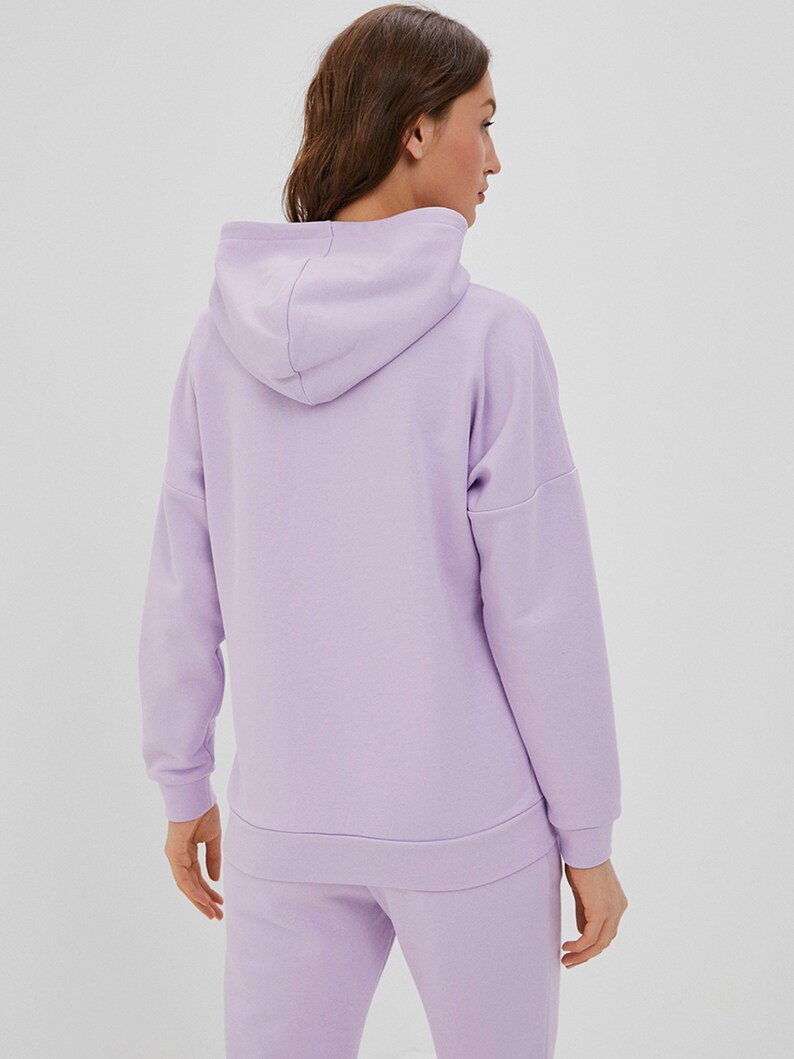 May include: A light purple hooded sweatshirt featuring long sleeves with ribbed cuffs, a matching ribbed hem, and a hood. The garment is constructed from a solid-colored fabric and is shown from the back, displaying the hood and sleeve seams.