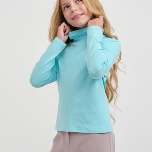 Può includere: Una maglia a maniche lunghe color azzurro con collo alto e vestibilità aderente. Il capo presenta una struttura in tessuto liscio a tinta unita con cuciture pulite lungo i polsini e l'orlo, offrendo un design semplice e funzionale.