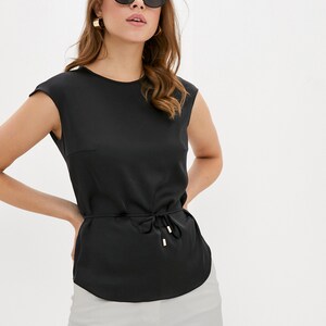 May include: A black sleeveless blouse featuring a round neckline, a cinched waist with a thin tie-front drawstring, and gold-toned metal aglets. The top has a smooth, satin-like finish and is paired with light gray trousers.