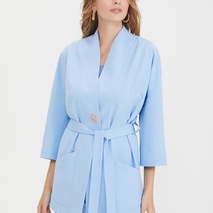 May include: A light blue blazer with three-quarter length sleeves, a shawl collar, and a matching fabric tie belt at the waist. The garment features a gold-toned decorative brooch shaped like a flower positioned at the center front closure.
