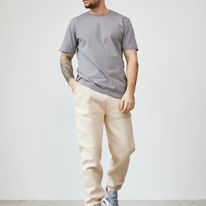 Puede incluir: Una camiseta gris lisa de manga corta con cuello redondo combinada con pantalones de chándal beige lisos con puños elásticos en los tobillos y zapatillas deportivas grises con suela blanca, formando un conjunto casual.