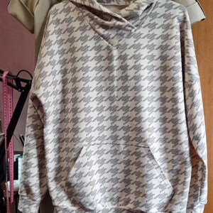 May include: A long-sleeved hooded sweatshirt featuring a repeating houndstooth pattern in shades of light beige and dark gray. The garment includes a front kangaroo pocket, a hood with a crossover neckline, and ribbed cuffs at the sleeves and hem.