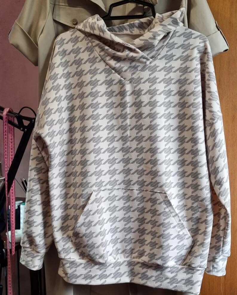 May include: A long-sleeved hooded sweatshirt featuring a repeating houndstooth pattern in shades of light beige and dark gray. The garment includes a front kangaroo pocket, a hood with a crossover neckline, and ribbed cuffs at the sleeves and hem.