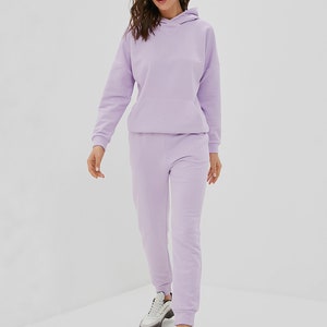 May include: A light purple matching set consisting of a long-sleeved hooded sweatshirt with a front kangaroo pocket and matching jogger-style sweatpants with ribbed cuffs, paired with white and grey athletic sneakers with thick soles.
