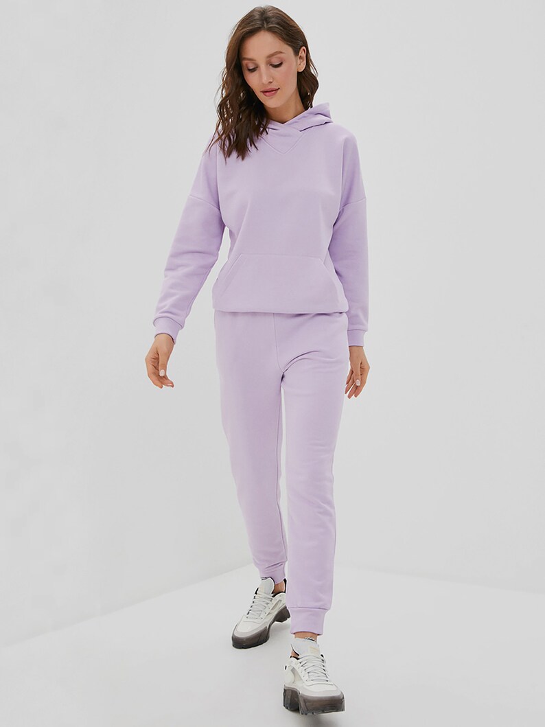 May include: A light purple matching set consisting of a long-sleeved hooded sweatshirt with a front kangaroo pocket and matching jogger-style sweatpants with ribbed cuffs, paired with white and grey athletic sneakers with thick soles.