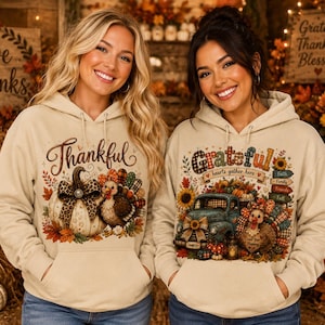 May include: Two beige hooded sweatshirts featuring Thanksgiving-themed graphics. The left hoodie displays a pumpkin with a leopard print bow and a turkey with the word Thankful. The right hoodie shows a blue truck, a turkey, and the word Grateful.