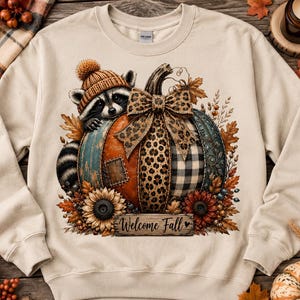 May include: A beige crewneck sweatshirt featuring a graphic of a raccoon wearing a knit hat, sitting behind a large pumpkin decorated with a leopard print bow, plaid patterns, and sunflowers. The text Welcome Fall is printed on a wooden sign.