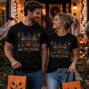 Pode incluir: T-shirts pretas com um design de Halloween. O design apresenta guaxinins com chapéus de bruxa, segurando uma abóbora, com o texto "Trick-Treat-Trash Repeat". O casal está segurando sacolas de mão laranja em forma de abóbora.