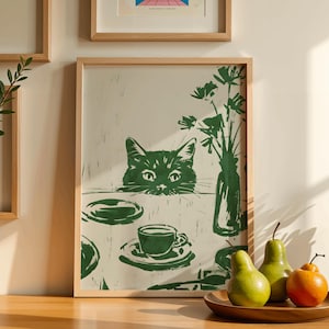 May include: Green and white framed art print featuring a cat peeking over a table set with a teacup, plates, and a vase of flowers. Two pears and an orange sit on a wooden plate in the foreground. The print has a minimalist style.