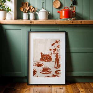 May include: Framed art print featuring a brown cat peeking over a table set with a cup, saucer, and vase of flowers. The artwork is in shades of brown and beige, with a black frame. The print is displayed in a kitchen setting.