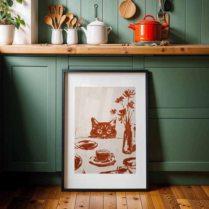 May include: Framed art print featuring a brown cat peeking over a table set with a cup, saucer, and vase of flowers. The artwork is in shades of brown and beige, with a black frame. The print is displayed in a kitchen setting.