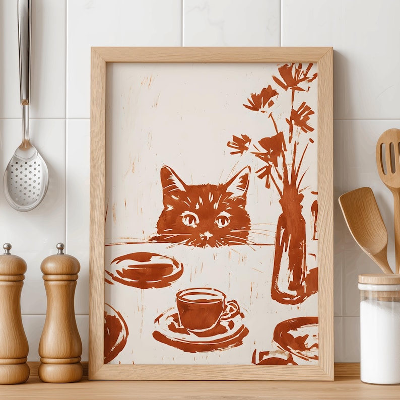 May include: A framed print featuring a brown cat peering over a table. The table is set with a cup of coffee, plates, and a vase of flowers. The artwork is in a warm, earthy color palette, creating a cozy, inviting feel. The frame is made of light-colored wood.
