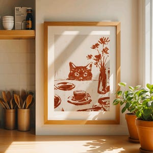 May include: A framed print featuring a cat peeking over a table set with a cup of coffee, plates, and a vase of flowers. The artwork is rendered in shades of brown and beige, creating a warm and inviting aesthetic. The print is displayed in a light wood frame.