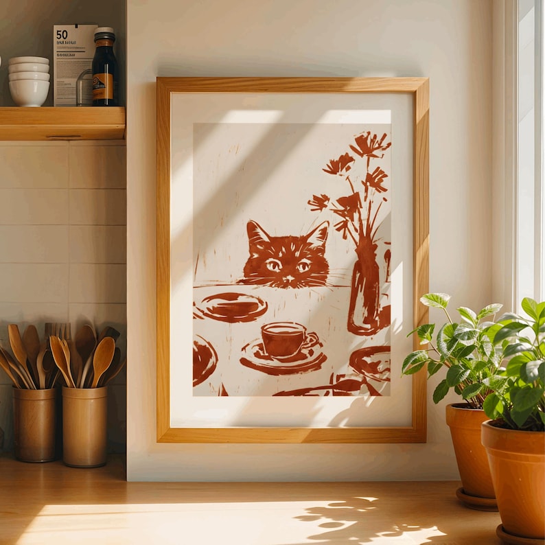 May include: A framed print featuring a cat peeking over a table set with a cup of coffee, plates, and a vase of flowers. The artwork is rendered in shades of brown and beige, creating a warm and inviting aesthetic. The print is displayed in a light wood frame.