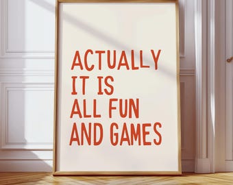 May include: A framed art print with the words "ACTUALLY IT IS ALL FUN AND GAMES" in red lettering. The print is set against a cream-colored background and framed in a light brown wooden frame. The artwork is leaning against a white wall.