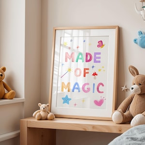 May include: A framed print with the words "MADE OF MAGIC" in colorful letters. The artwork includes illustrations of stars, a butterfly, a mushroom, and a heart. The frame is light brown, and the print is displayed on a wooden surface.