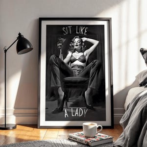 May include: Monochrome framed print with the text "SIT LIKE A LADY". The image shows a woman in a bra and trousers, seated in a chair, holding a cocktail. A black lamp, a coffee cup, and a book are in the foreground.