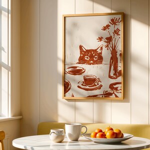 May include: A framed print featuring a cat peering over a table set with dishes, a teacup, and a vase of flowers. The artwork is in shades of brown and cream, with a rustic, linocut style. The print is displayed in a room with a round table and a plate of oranges.