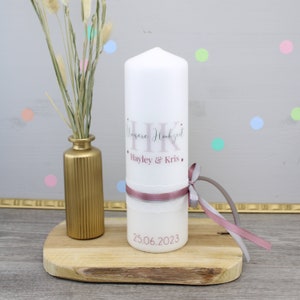 Könnte beinhalten: Weiße Hochzeitskerze mit rosa Schleife und Spitzenbesatz. Die Kerze ist mit dem Text "Unsere Hochzeit" und den Namen "Hayley & Kris" in rosa Schrift verziert. Das Datum "25.06.2023" ist auf der Unterseite der Kerze aufgedruckt.