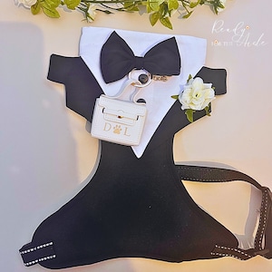 May include: A black dog harness with a white tuxedo-style collar, a black bow tie, a white purse with the letters "D&L" and a paw print, and a white flower.