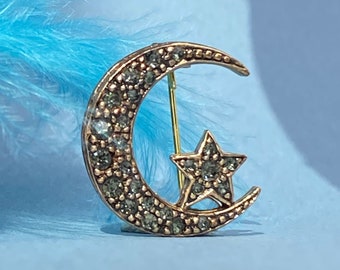 May include: A gold-toned crescent moon and star brooch with clear rhinestones. The brooch is on a light blue background.