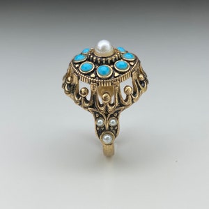 May include: A gold ring featuring a large, round, turquoise-coloured stone encircled by smaller turquoise stones. The ring is adorned with a pearl-like centre stone and a crown-like design.