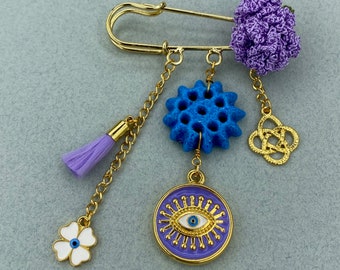 May include: A gold safety pin with a purple crocheted flower charm, a blue and gold charm, a gold knot charm, a purple tassel, and a white flower charm with a blue evil eye charm.