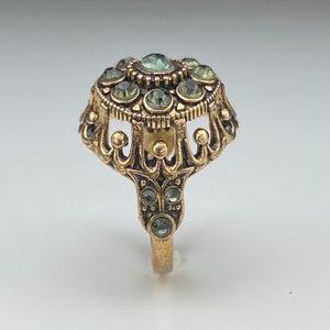 Op de afbeelding: Een gouden ring met een kroonvormig ontwerp en een cluster van groene strass-steentjes in het midden.