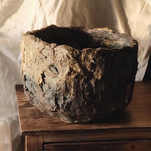 Può includere: Un vaso da fiori in ceramica rustico, marrone e blu con una superficie ruvida e testurizzata. Il vaso è appoggiato su un tavolo di legno.