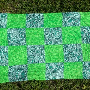 May include: A rectangular quilt with a patchwork design. The quilt features alternating squares of solid bright green and patterned green fabric. The patterned squares have a swirling design in white and teal. The quilt is lying on green grass.