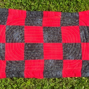 May include: A rectangular, quilted mat with a checkerboard pattern of red and dark blue fabric. The dark blue squares feature a floral print. Gold stitching outlines a curved design in the centre. Metal eyelets are in each corner.