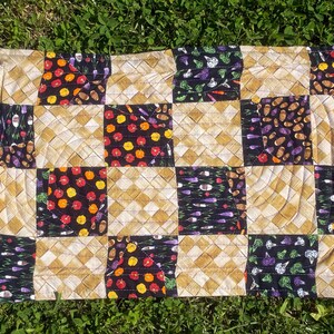 May include: Rectangular quilted placemat with a patchwork design. The squares feature alternating patterns of woven tan fabric and black fabric with colourful illustrations of vegetables and fruits, including tomatoes, carrots, and broccoli.