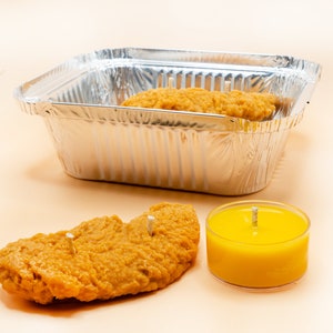 Several chicken tender shaped candles are placed inside a aluminum foil box. One piece of candle and a yellow mustard tea light are placed in the front.