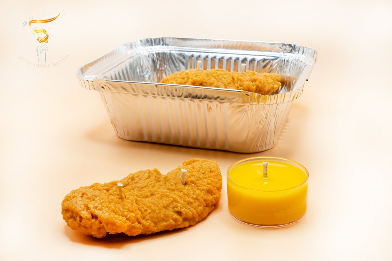 Several chicken tender shaped candles are placed inside a aluminum foil box. One piece of candle and a yellow mustard tea light are placed in the front.