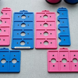 May include: A collection of rectangular 3D printed storage organisers in bright blue and pink. The organisers feature circular cutouts and small holes, with one blue piece displaying the text DESIGNED & PRINTED IN UK. These modular units are arranged on a flat surface.