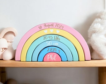 May include: A rainbow-colored wooden arch with five rings, decorated with a baby's birth details. The top ring is pink and says "18 August 2024" with three white hearts. The next ring is yellow and says "20:45 Uhr". The third ring is light blue and says "50 cm". The fourth ring is blue and says "2870 g". The bottom ring is pink and says "Phil".