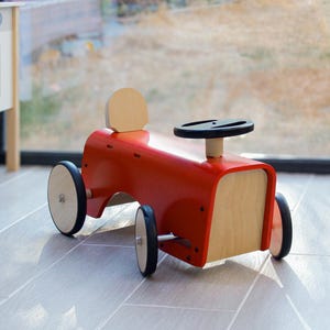 Peut inclure: Une voiture jouet à pousser en bois rouge avec un panneau avant et un dossier de siège en bois clair naturel. Le véhicule est équipé de quatre roues avec des pneus en caoutchouc noir, d'un volant noir et d'un design épuré en bois lisse.