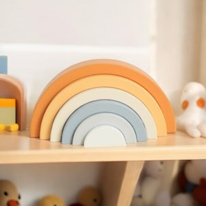 Puede incluir: Un juguete de apilar arcoíris de madera con cinco arcos en tonos naranja, amarillo, crema, azul y blanco.