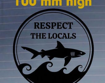 May include: A black circular decal featuring a shark silhouette swimming above stylized waves. The text "RESPECT THE LOCALS" is printed in black capital letters inside the circle. The decal is clearly marked as being 100 mm high.