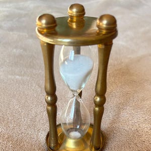 Könnte beinhalten: Eine Sanduhr mit einem goldfarbenen Metallrahmen, drei vertikalen Säulen sowie einem runden Sockel und Deckel. Der klare Glaskörper enthält weißen Sand, wobei sich ein Teil des Sandes derzeit in der unteren Kammer des Glasgefäßes befindet.