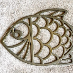 Puede incluir: Un salvamanteles decorativo con forma de pez, hecho de metal. El salvamanteles es de color gris metálico apagado y presenta un diseño calado con elementos circulares y curvos. El pez tiene cola y aletas detalladas. Mide aproximadamente 25 cm de largo.