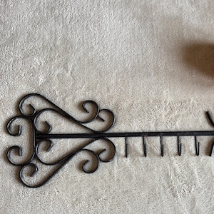 Puede incluir: Perchero de llaves de metal negro con adornos decorativos y siete ganchos. Dos peonías rosas con tallos y hojas verdes en el fondo. El perchero de llaves es un elemento decorativo para el hogar.