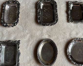 May include: A collection of seven silver-toned metal trays in various shapes, including rectangular and oval designs. Each tray features decorative embossed borders and smooth, reflective centers, with some displaying subtle engraved crests or markings.