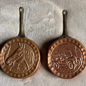 Puede incluir: Dos sartenes de color cobre con mangos largos. Una sartén presenta una cabeza de caballo, la otra un coche antiguo. Las sartenes tienen una textura martillada y un lazo al final del mango para colgar.