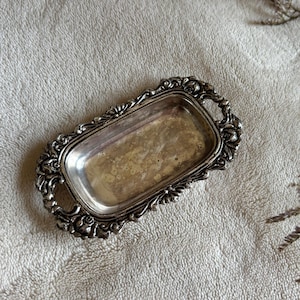 May include: A rectangular silver-toned metal tray featuring ornate, raised floral and scrollwork detailing along the edges and integrated handles on both ends. The central surface of the tray has a slightly tarnished, weathered metallic finish.