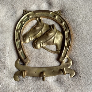 Op de afbeelding: Een messing wandhaakrek met een hoefijzervormig frame en twee paardenkoppen in reliëf in het midden. De onderkant van het hoefijzer loopt uit in een horizontale balk met drie kleine, gebogen haken voor het ophangen van voorwerpen, goudkleurig.