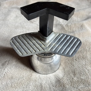 Puede incluir: Una cafetera espresso plateada con un asa negra. La base es cilíndrica y el asa tiene forma de T. La cafetera tiene un componente acanalado en forma de ala. El texto "Nova Espresso" es visible en la base.