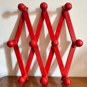 May include: A red wooden accordion-style wall-mounted coat rack. The rack has a diamond-shaped design with red wooden knobs at each intersection. The rack is displayed on a wooden surface against a white wall.